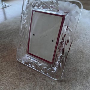 Könnte beinhalten: Klarer Glas-Fotorahmen mit geätzten Blumen- und Blattmotiven. Der Rahmen hat eine rechteckige Öffnung mit einem bordeauxfarbenen Rand. Der Rahmen liegt auf einer strukturierten, hellbraunen Oberfläche.