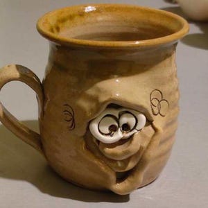 Può includere: Una tazza in ceramica marrone con un viso fantasioso scolpito sul davanti. La tazza ha un manico grande e una finitura smaltata.