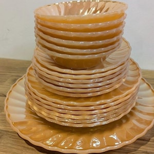 Puede incluir: Un conjunto de vajilla de vidrio color melocotón, que incluye cuencos y platos. El conjunto vintage presenta un diseño de borde festoneado. El vidrio tiene una calidad iridiscente, creando un brillo cálido. Los platos están apilados, destacando su elegante forma. Medidas en centímetros.