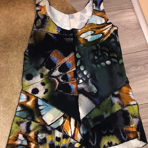 Puede incluir: Una camiseta sin mangas con fondo negro y estampado de mariposas de colores. El estampado presenta tonos de azul, verde, marrón y amarillo.