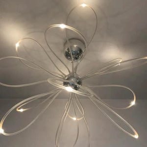 Op de afbeelding: Een moderne plafondlamp met een bloemvormig ontwerp. De lamp heeft een centrale chromen bol met meerdere gebogen, witte, verlichte armen die naar buiten uitsteken, elk met een klein licht aan het uiteinde. De lamp is gemonteerd op een wit plafond.