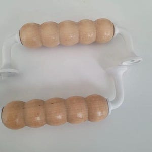 May include: Two white cabinet handles with light brown, cylindrical wooden grips. The handles have a curved design and are mounted on a white surface. The wooden grips have a smooth, natural finish.