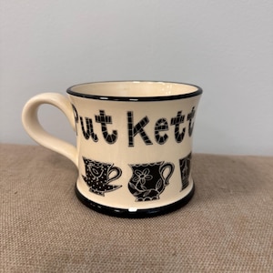 Puede incluir: Taza de cerámica color crema con borde y asa negros. La taza presenta las palabras "Put kett" en letras negras y ilustraciones de tazas de té negras. La taza está sobre una superficie marrón.