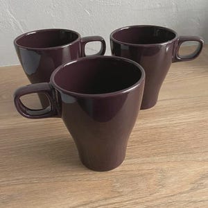 May include: Three plum-colored ceramic mugs with rounded handles are arranged on a light wood-grain surface. The mugs have a tapered shape, wider at the top and narrowing towards the base. The mugs are empty.