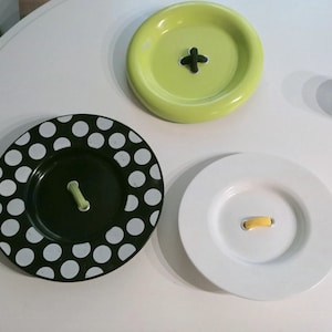 May include: Three decorative plates with a button design. The plates are white, black with white polka dots, and green. Each plate has a different button design.