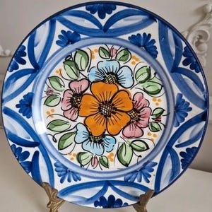 Puede incluir: Un plato de cerámica pintado a mano con un diseño floral. El plato presenta una flor naranja central rodeada de flores rosas y azules, hojas verdes y un borde azul y blanco. El plato se exhibe sobre un soporte dorado.