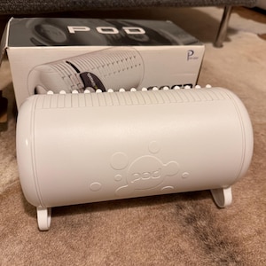 May include: A white, cylindrical office product with the word "POD" printed on the front. The product has a series of small, raised ridges along the top and is supported by two small legs. A box with the word "POD" is in the background.