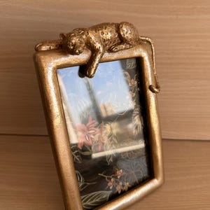 May include: A gold-coloured picture frame with a decorative leopard resting on the top. The frame has a rectangular shape and features a floral design inside. The frame is made of a metallic material.