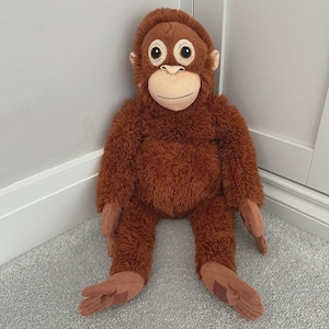 May include: A plush orangutan toy with brown fur, large round eyes, and a tan face. The stuffed animal is sitting upright with its arms and legs extended. The toy is positioned against a light gray wall and a white door frame.