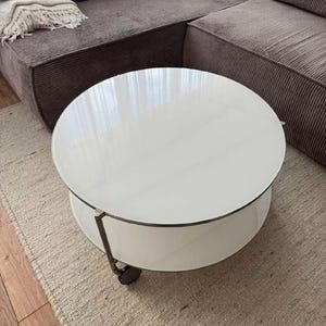 Puede incluir: Una mesa de centro redonda y moderna con una superficie blanca y un estante inferior blanco. La mesa tiene una estructura metálica y pequeñas ruedas. La mesa tiene un diámetro de aproximadamente 91 cm.