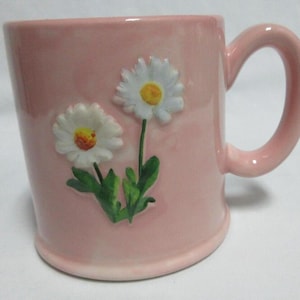 May include: A pink ceramic mug with a curved handle. The mug features two white daisy flower designs with yellow centers and green stems. The mug is a light pink color.