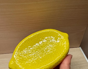 Könnte beinhalten: Eine hellgelbe Keramikschale in Zitronenform. Die Schale hat eine strukturierte Oberfläche und ein glänzendes Finish. Die zitronenförmige Schale wird in einer Hand gehalten.