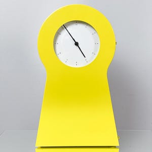 May include: A bright yellow table clock with a smooth, curved, arch-shaped frame. The clock face is circular and white, featuring black numerals from 1 to 12 and two thin black hands indicating the time. The design is solid and uniform in color.