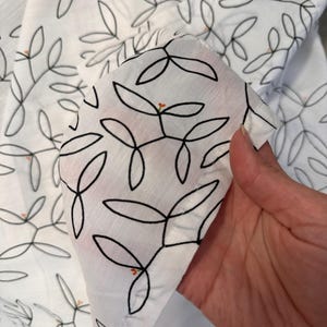 May include: White fabric featuring a black leaf pattern with small orange accents. The fabric is held up, showcasing the detailed design. The leaves are outlined in black, creating a delicate look. The overall theme is a botanical or nature-inspired textile.