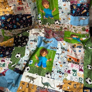 Op de afbeelding: Een patchworkquilt met diverse stofpatronen. De quilt heeft vierkanten met voetballen, vliegtuigen, dinosaurussen en een stripfiguur. Andere vierkanten hebben bouwmachines, katten en auto's. Het kleurenpalet omvat blauw, groen, geel en zwart.