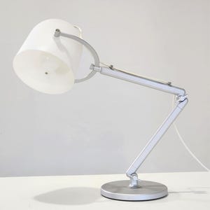 Peut inclure: Une lampe de bureau argentée avec un abat-jour blanc et un bras flexible. La lampe est posée sur une surface blanche.