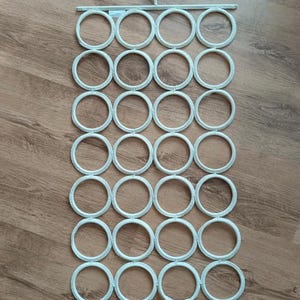 May include: A white scarf organizer with a hook for hanging. The organizer has a rectangular frame with 28 evenly spaced circular rings. The rings are designed to hold scarves, belts, or other accessories. The organizer is made of a lightweight material.