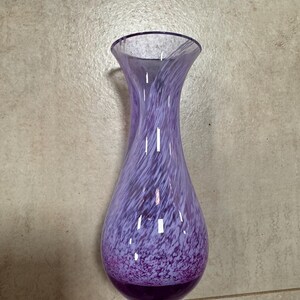 Puede incluir: Un jarrón de vidrio morado con motas blancas y un diseño de espiral. El jarrón tiene un cuello estrecho y un fondo redondeado.