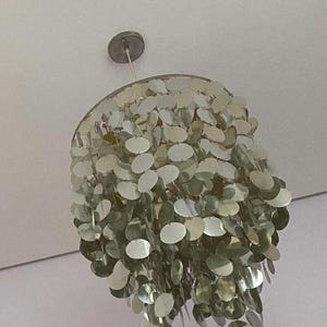 May include: A silver-toned chandelier with a multitude of overlapping, oval-shaped, reflective discs. The light fixture is suspended from a circular ceiling mount. The discs create a layered, textured effect, reflecting light.