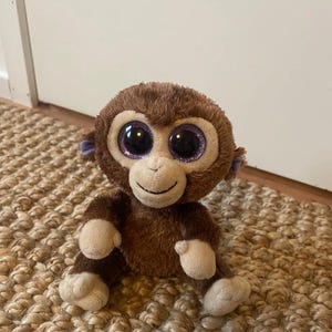 Peut inclure: Une peluche singe marron avec de grands yeux violets scintillants et des mains et des pieds beiges. L'animal en peluche a un visage souriant et de petites oreilles violettes. Le singe est assis sur un tapis tissé.