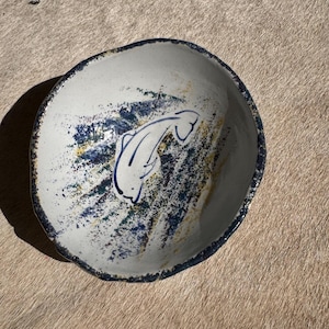 May include: A handmade ceramic bowl with a white interior and a dark blue rim. The bowl features a blue fish design and blue and yellow paint splatters. The bowl has an irregular, organic shape.