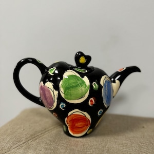 Pode incluir: Um bule de cerâmica preta com um design pintado à mão de grandes pontos circulares coloridos em tons de verde, laranja, azul e roxo. O bule possui uma alça curva, um bico e uma tampa com um pequeno puxador em formato de coração.