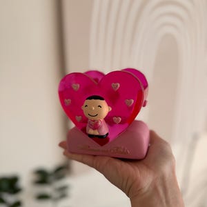 May include: Pink heart-shaped figurine with a small cartoon character holding a heart inside. The figurine has the words "Love you lots" written on the base.