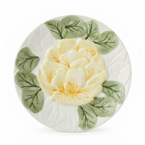 May include: A round white ceramic plate featuring a raised, three-dimensional yellow flower at the center. The flower is surrounded by embossed green leaves with dark green veining. The plate surface has a subtle, molded leaf pattern texture.