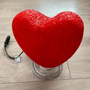 May include: A red heart-shaped lamp with a textured surface sits on a silver spring base. The lamp is plugged in and ready to be turned on.
