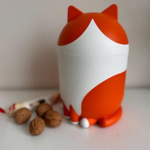 Puede incluir: Un recipiente caprichoso con forma de zorro, naranja y blanco. El recipiente tiene una franja blanca en el medio y orejas de zorro. Varias nueces y un pequeño caramelo envuelto están junto al recipiente.