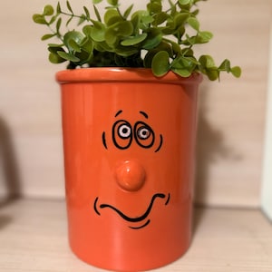 Puede incluir: Una maceta de cerámica naranja con un diseño de cara de dibujos animados, con ojos negros, una nariz y una boca sonriente. La maceta está llena de follaje artificial verde, creando una pieza decorativa caprichosa.