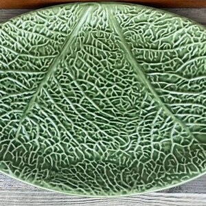 Peut inclure: Un plat ovale en céramique de couleur vert clair, présentant une texture en relief détaillée qui imite les motifs complexes des nervures et la structure organique d'une grande feuille de chou, couvrant toute la surface de l'objet.