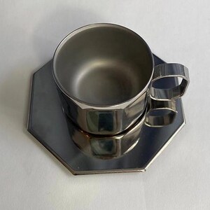 Puede incluir: Un juego de taza y platillo de acero inoxidable. La taza tiene un dise&ntilde;o geom&eacute;trico y un asa cuadrada. El platillo tiene una forma a juego. El juego es reflectante y se encuentra sobre una superficie blanca.