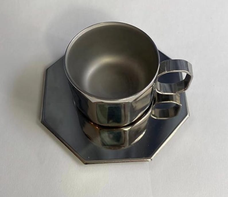 Puede incluir: Un juego de taza y platillo de acero inoxidable. La taza tiene un dise&ntilde;o geom&eacute;trico y un asa cuadrada. El platillo tiene una forma a juego. El juego es reflectante y se encuentra sobre una superficie blanca.