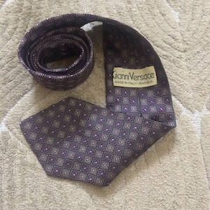 Puede incluir: Una corbata de seda morada con un patrón geométrico y una etiqueta que dice "Gianni Versace Made in Italy - 100% Silk."