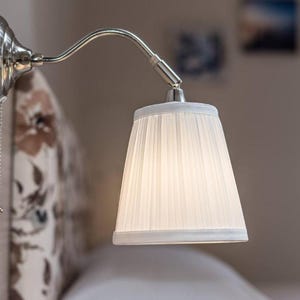 Può includere: Una lampada da comodino a parete con finitura argentata. La lampada presenta un braccio curvo e un paralume bianco plissettato. Una piccola catena pende per l'accensione e lo spegnimento. La lampada è montata su una parete chiara.