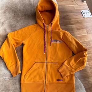 Puede incluir: Una sudadera con capucha de color amarillo mostaza con un cordón morado y el texto "Peak Performance" en una fuente colorida.