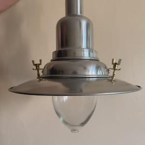 Puede incluir: Una lámpara colgante de estilo vintage con acabado metálico cepillado. Tiene una parte superior redondeada, una pantalla ancha y plana y una carcasa de vidrio transparente en forma de lágrima. Pequeños detalles de latón acentúan la pantalla. La lámpara está suspendida de una varilla metálica.