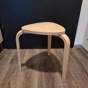Könnte beinhalten: Ein dreibeiniger Hocker aus hellem Holz mit drei Beinen. Der Hocker hat eine glatte, abgerundete Oberseite.