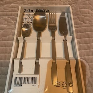 Könnte beinhalten: Ein verpacktes Set aus Edelstahlbesteck, einschließlich Löffeln, Gabeln und Messern. Die Verpackung trägt die Aufschrift "24x DATA" und enthält den Designnamen und die Produktnummer. Das Set ist in einem weißen Kartoneinsatz angeordnet.