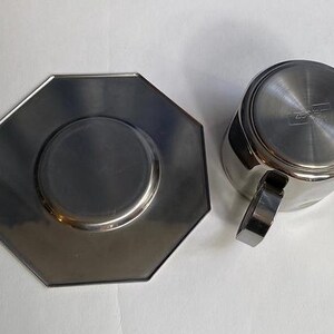 Puede incluir: Juego de taza y platillo de caf&eacute; de acero inoxidable. La taza tiene un asa y forma cil&iacute;ndrica. El platillo es octogonal con un centro rebajado. Ambas piezas tienen un acabado met&aacute;lico brillante.
