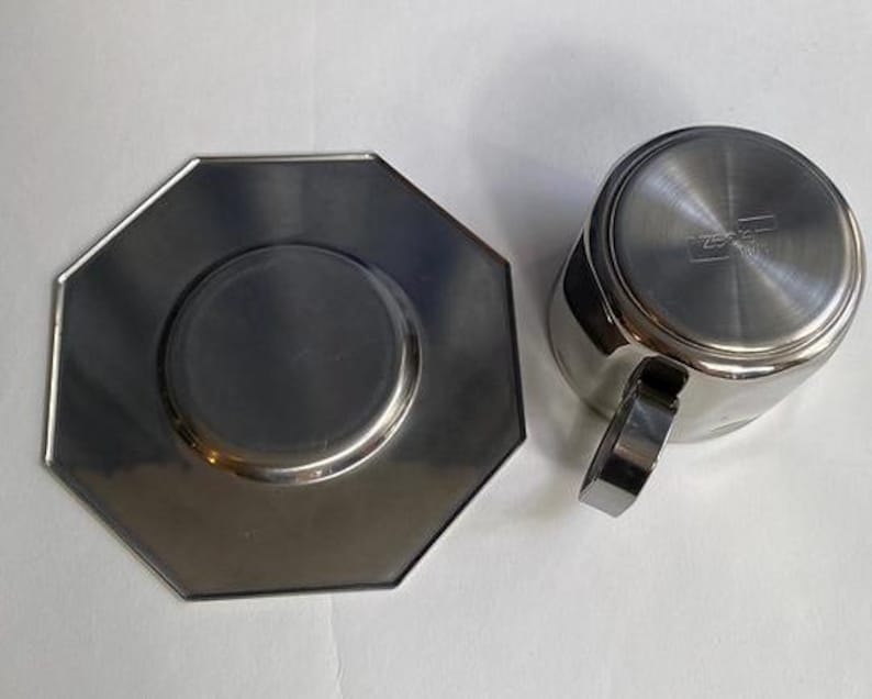 Puede incluir: Juego de taza y platillo de caf&eacute; de acero inoxidable. La taza tiene un asa y forma cil&iacute;ndrica. El platillo es octogonal con un centro rebajado. Ambas piezas tienen un acabado met&aacute;lico brillante.