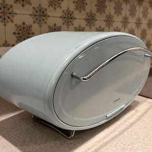 May include: A pale blue, oval bread bin with a chrome handle and base. The bread bin has a curved design and a hinged lid. The word "Tramontina" is printed on the front of the bread bin.