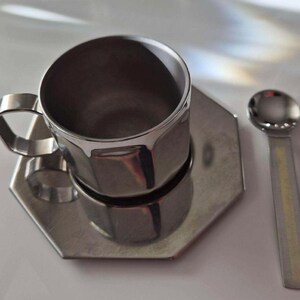 Puede incluir: Un juego de taza de caf&eacute;, platillo y cuchara de acero inoxidable. La taza tiene un dise&ntilde;o geom&eacute;trico y un asa. El platillo es hexagonal. La cuchara tiene un cuenco peque&ntilde;o y redondo y un mango plano. El juego es de metal brillante color plata.