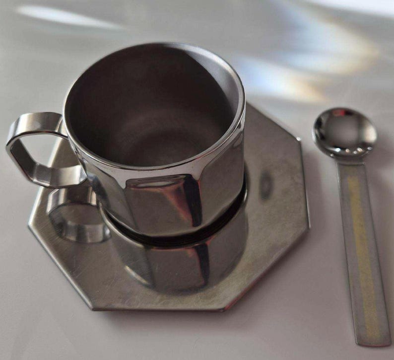 Puede incluir: Un juego de taza de caf&eacute;, platillo y cuchara de acero inoxidable. La taza tiene un dise&ntilde;o geom&eacute;trico y un asa. El platillo es hexagonal. La cuchara tiene un cuenco peque&ntilde;o y redondo y un mango plano. El juego es de metal brillante color plata.
