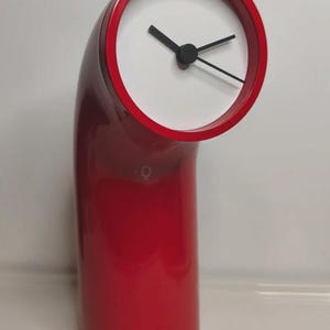 May include: A glossy red table clock with a curved, tubular body and a circular face. The clock face is white with two black hands, positioned at the top of the angled structure. The design features a smooth, solid red finish throughout the base.
