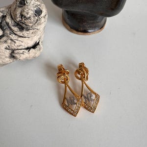 May include: A pair of gold-tone dangle earrings with a diamond-shaped pendant featuring the letters "M.N.C." in white metal. The earrings have a delicate chain design and are adorned with clear rhinestones.