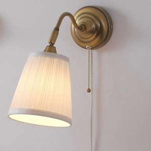 Può includere: Applique da parete in ottone con paralume bianco plissettato. La lampada ha una catena e un design per uso interno. L'apparecchio è montato su una parete bianca. Ideale per l'illuminazione d'ambiente.