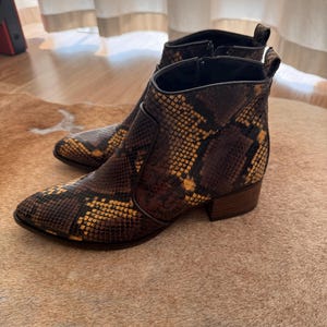 May include: Ankle boots featuring a snakeskin design in brown and yellow hues. These boots have a pointed toe, a low heel, and a zip closure. The textured material creates a stylish look. The boots are a fashionable footwear choice.