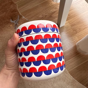 Puede incluir: Tarro de cerámica blanco con tapa, decorado con un patrón repetitivo de semicírculos rojos y azules. El tarro es cilíndrico y parece hecho a mano. El diseño recuerda a un estilo retro o moderno de mediados de siglo.
